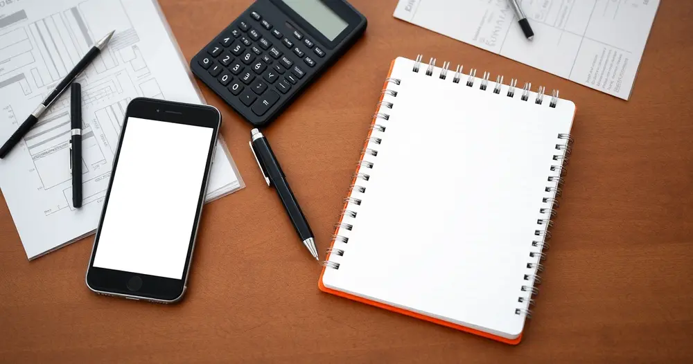 Caderno com páginas em branco, caneta e calculadora sobre mesa ao lado de celular
